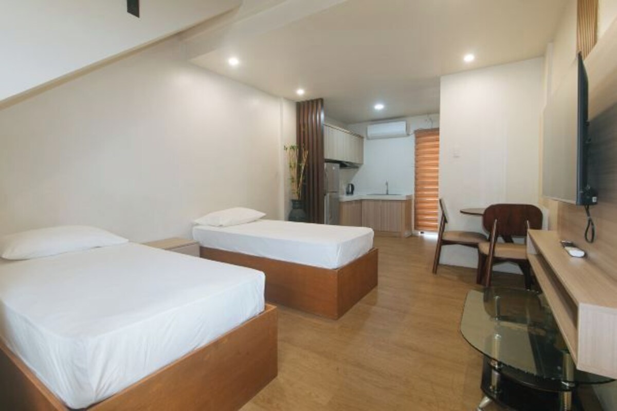 A hotel room with two beds, a small kitchenette, a dining area with a glass table, and a TV mounted on the wall.