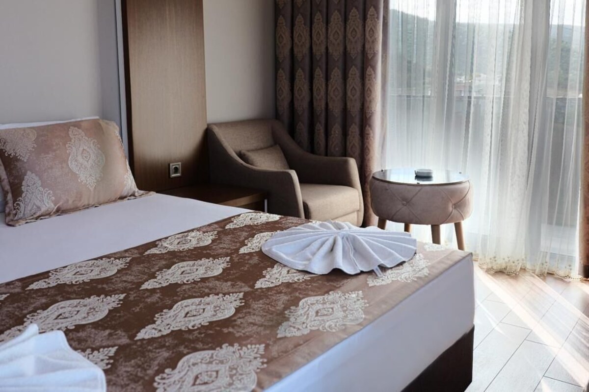 A hotel room with a large bed, a sitting area with a chair and a small table, and a view through sheer curtains.