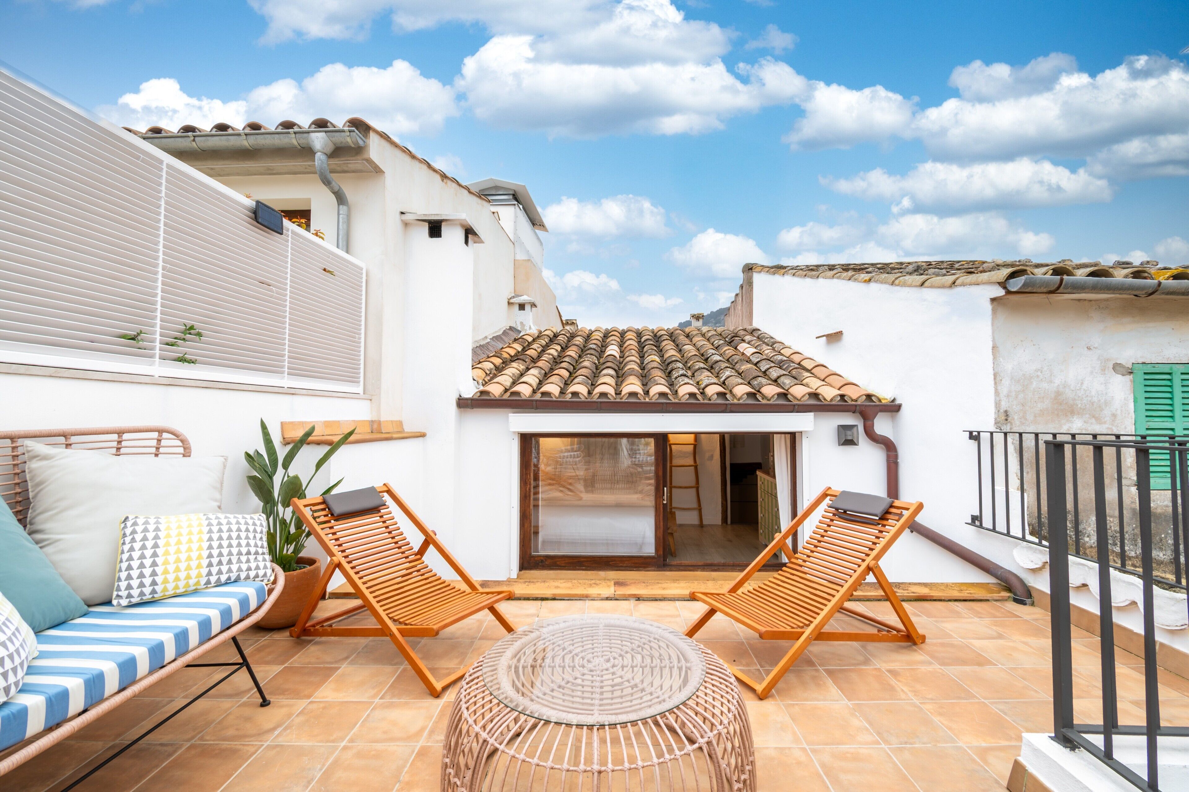 Eine Terrasse mit zwei Holzliegestühlen, einem runden gewebten Hocker, einer gepolsterten Couch mit gemusterten Kissen und einem Blumentopf.
