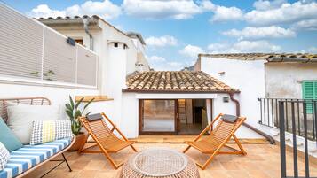 Eine Terrasse mit zwei Holzliegestühlen, einem runden gewebten Hocker, einer gepolsterten Couch mit gemusterten Kissen und einem Blumentopf.