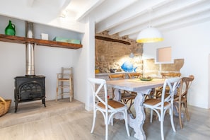 Un coin repas agrémenté d’un mur en pierre rustique, d’une table en bois, de chaises blanches et d’un poêle noir.