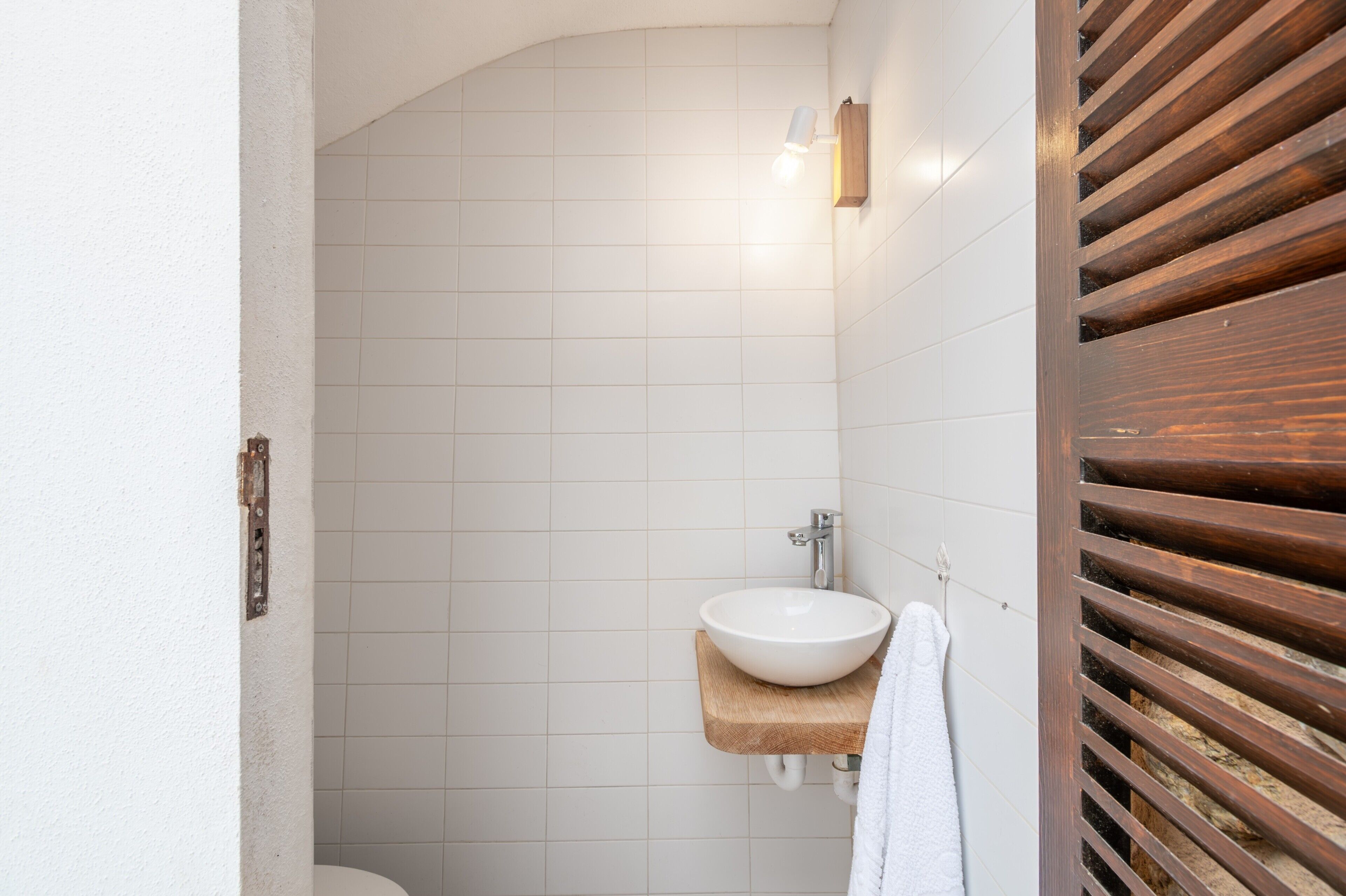 Un baño con un lavamanos blanco sobre una repisa de madera, paredes de azulejos blancos y una puerta de madera con listones horizontales.