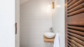 Un baño con un lavamanos blanco sobre una repisa de madera, paredes de azulejos blancos y una puerta de madera con listones horizontales.