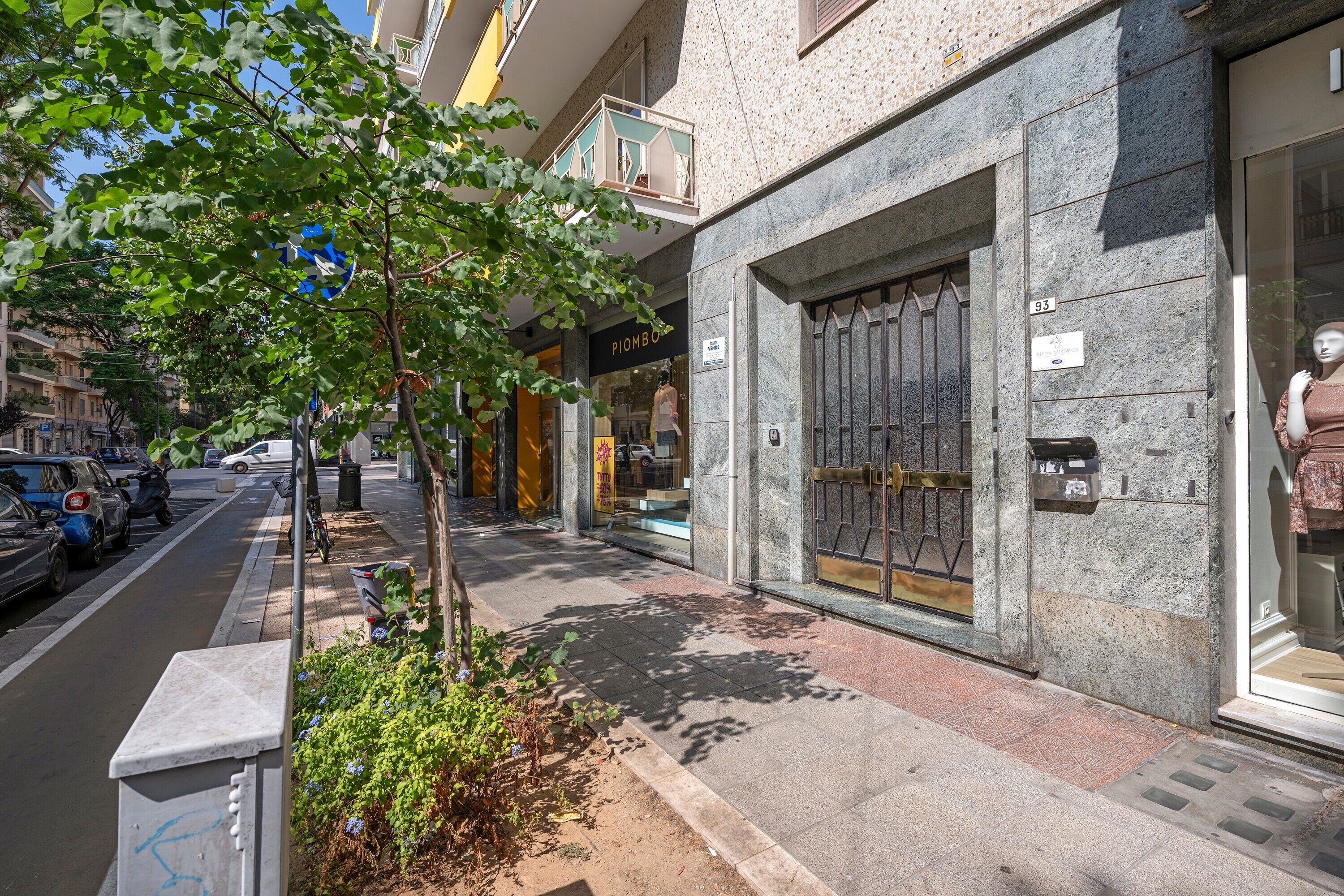 A street view with a building featuring a large glass door, a sign indicating a sale, and a tree in the foreground.