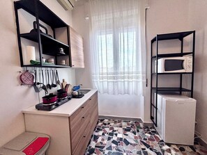 Una cocina compacta con microondas, refrigerador, piso de baldosas y ventana con cortinas translúcidas.