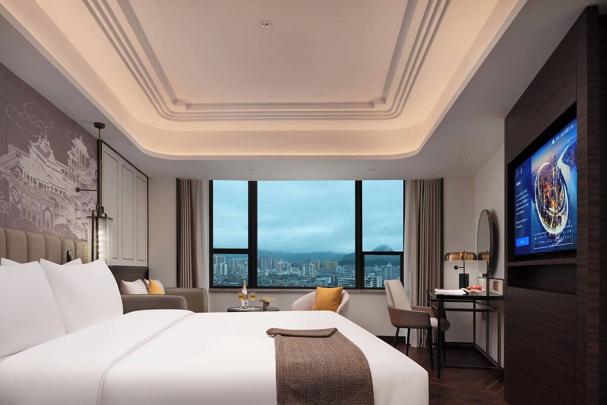 Ein modernes Hotelzimmer mit einem großen Bett, einem Schreibtisch, einem Fernseher und Blick auf die Stadt.