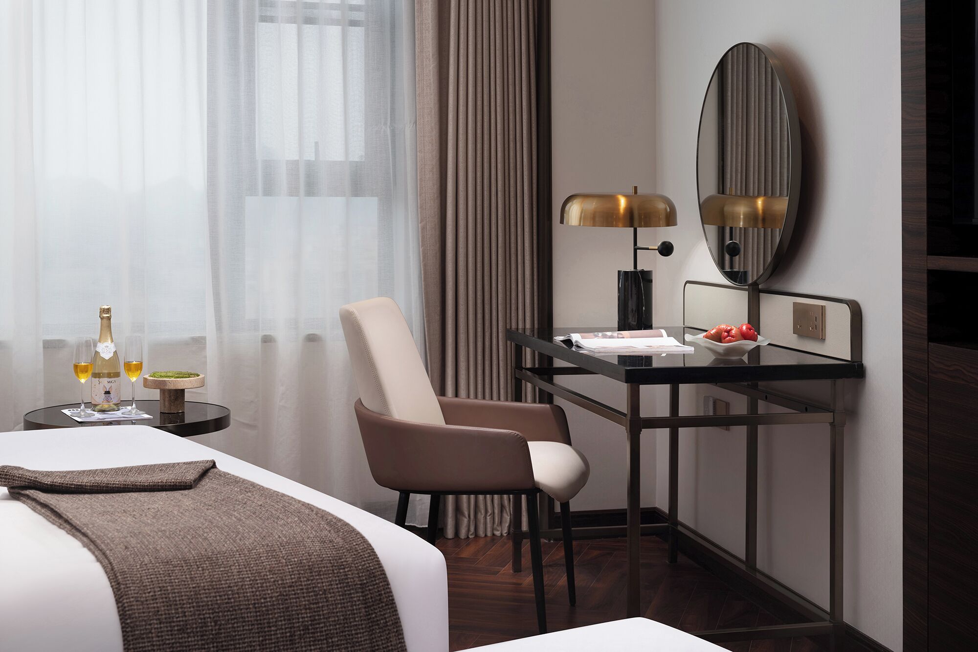 Habitación de hotel con cama, escritorio con lámpara, silla y una pequeña mesa con una botella y vasos.