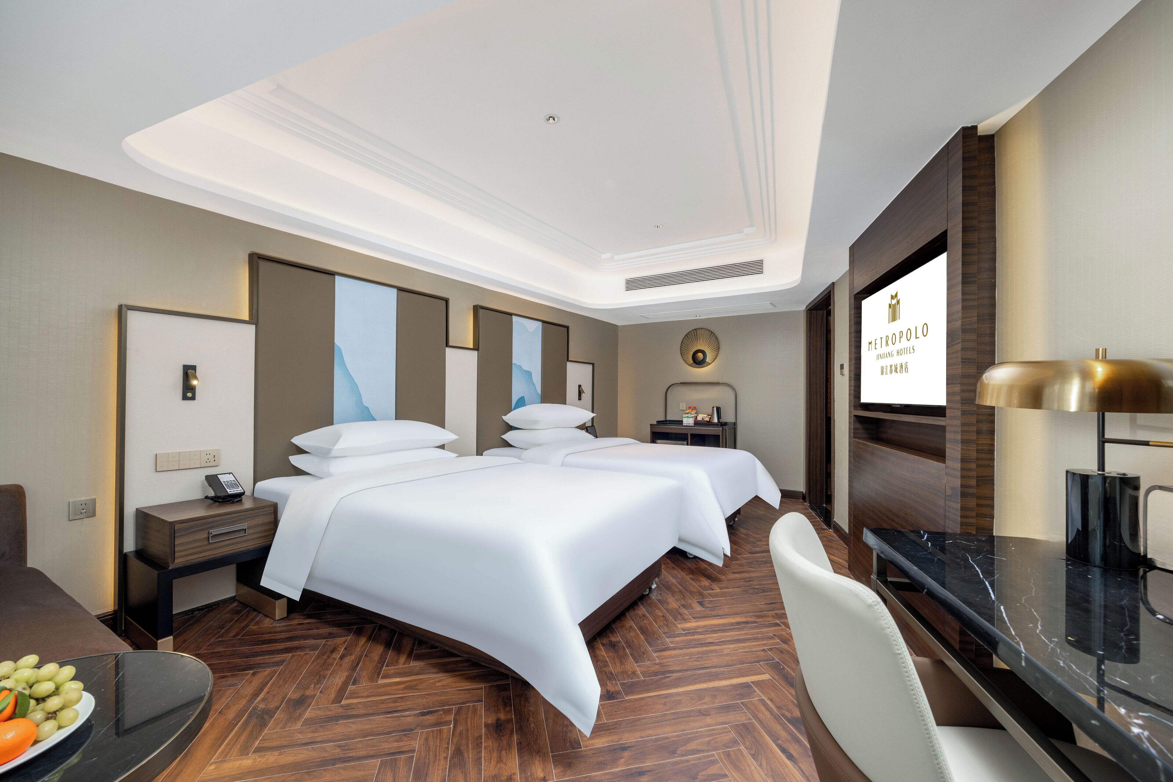 A hotel room with two beds, a desk, a chair, a dining table with fruit, and a TV screen displaying hotel branding.