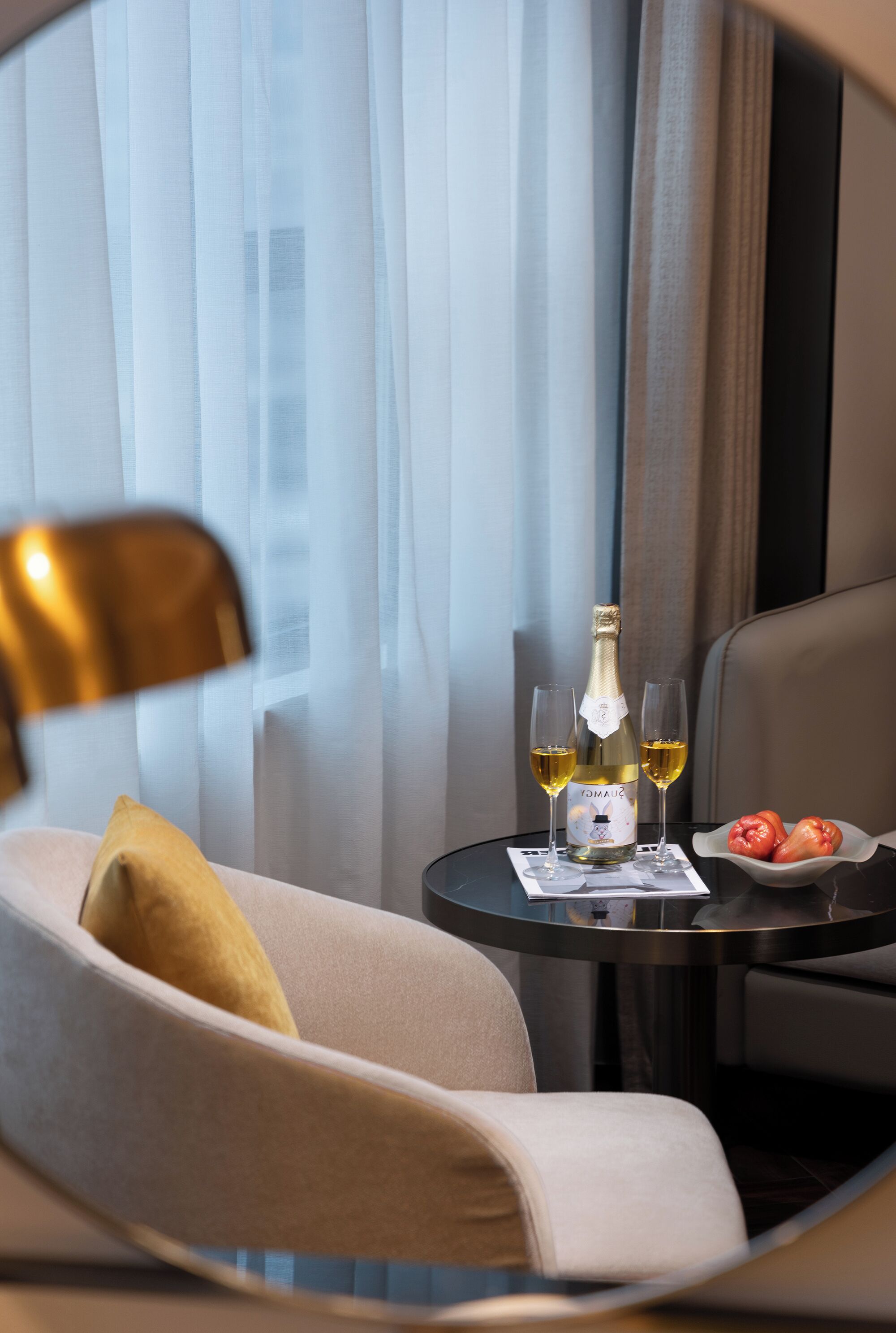A hotel room with a bed, a small table with a bottle of champagne, two glasses, and a bowl of fruit.
