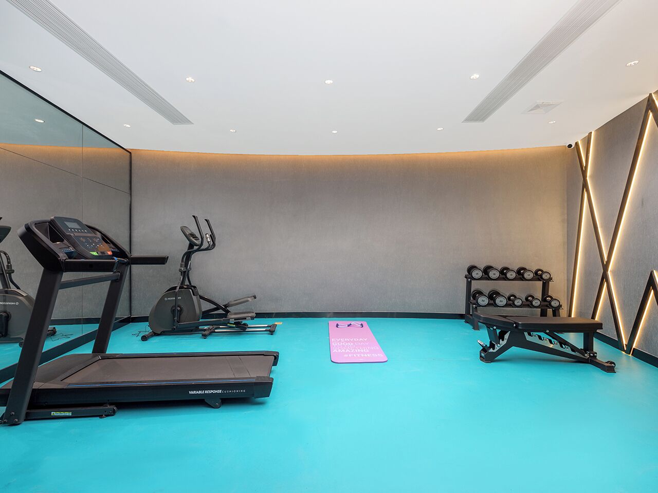 Un gimnasio moderno con suelo azul, cintas de correr, elíptica, banco de pesas y una barra con mancuernas.