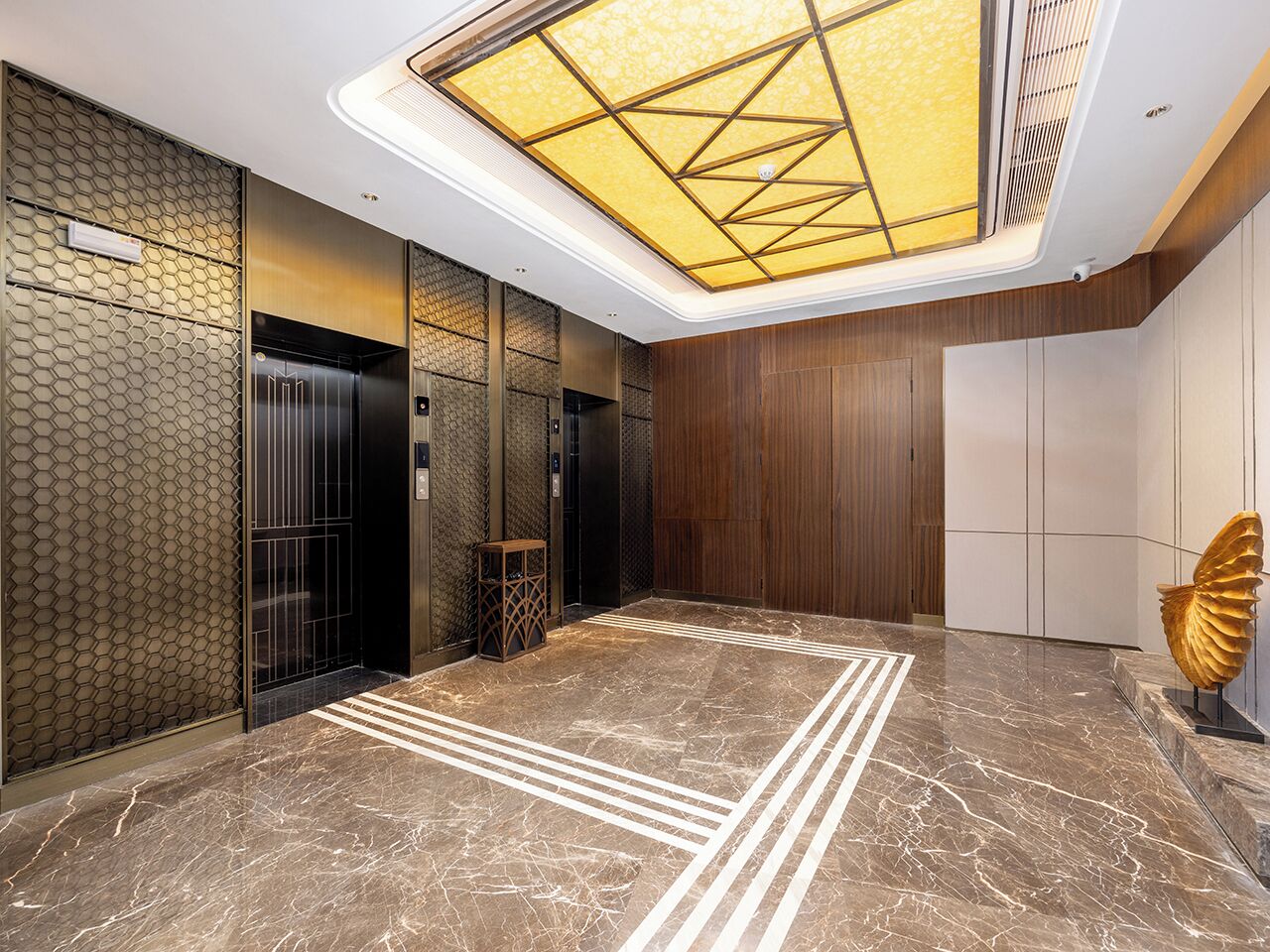 A modern elevator area with a geometric ceiling design, marble flooring, and a decorative golden sculpture.