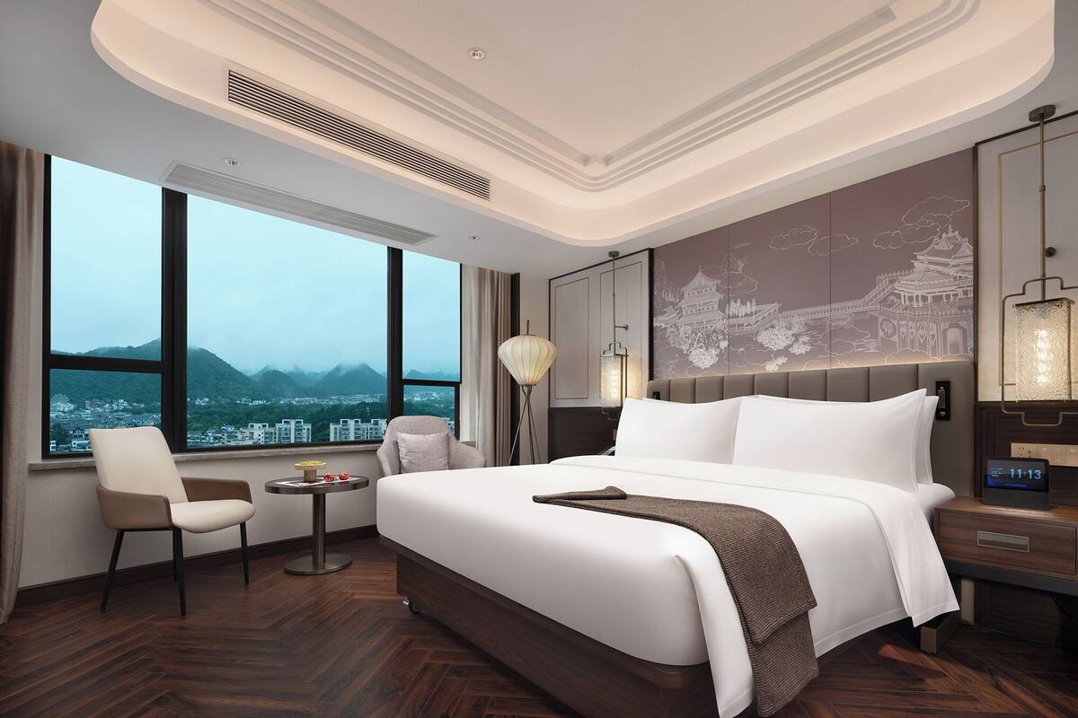 Ein modernes Hotelzimmer mit einem großen Bett, einem Sessel, einem kleinen Tisch und Blick auf eine Stadtlandschaft durch große Fenster.