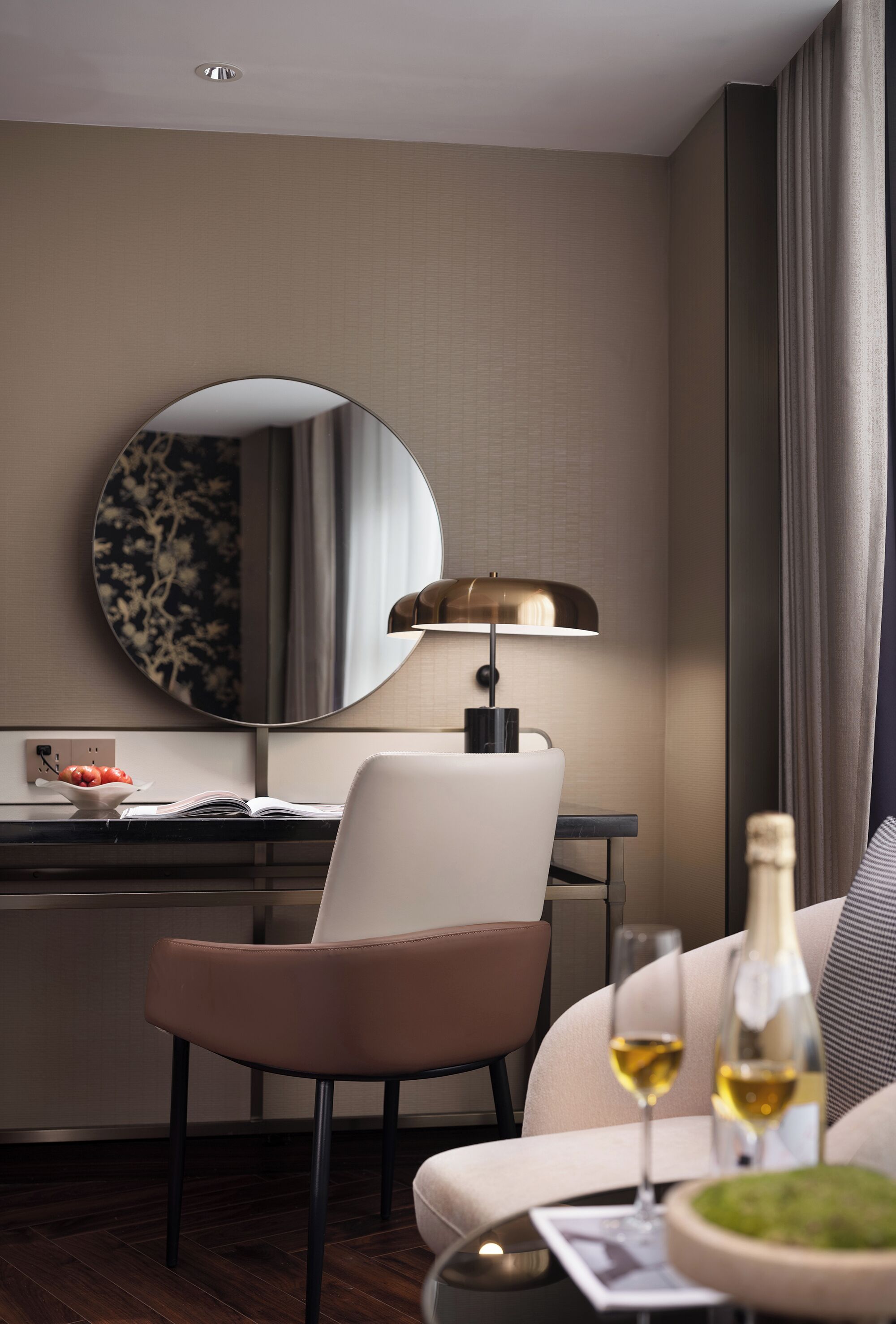 Een moderne hotelkamer met een ronde spiegel, een bureau met een laptop, een comfortabele bank, een klein tafeltje met een fles en glazen, en een decoratieve plant.