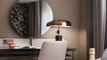 Une chambre d’hôtel moderne avec un miroir rond, un bureau avec un ordinateur portable, un canapé confortable, une petite table avec une bouteille et des verres, et une plante décorative.