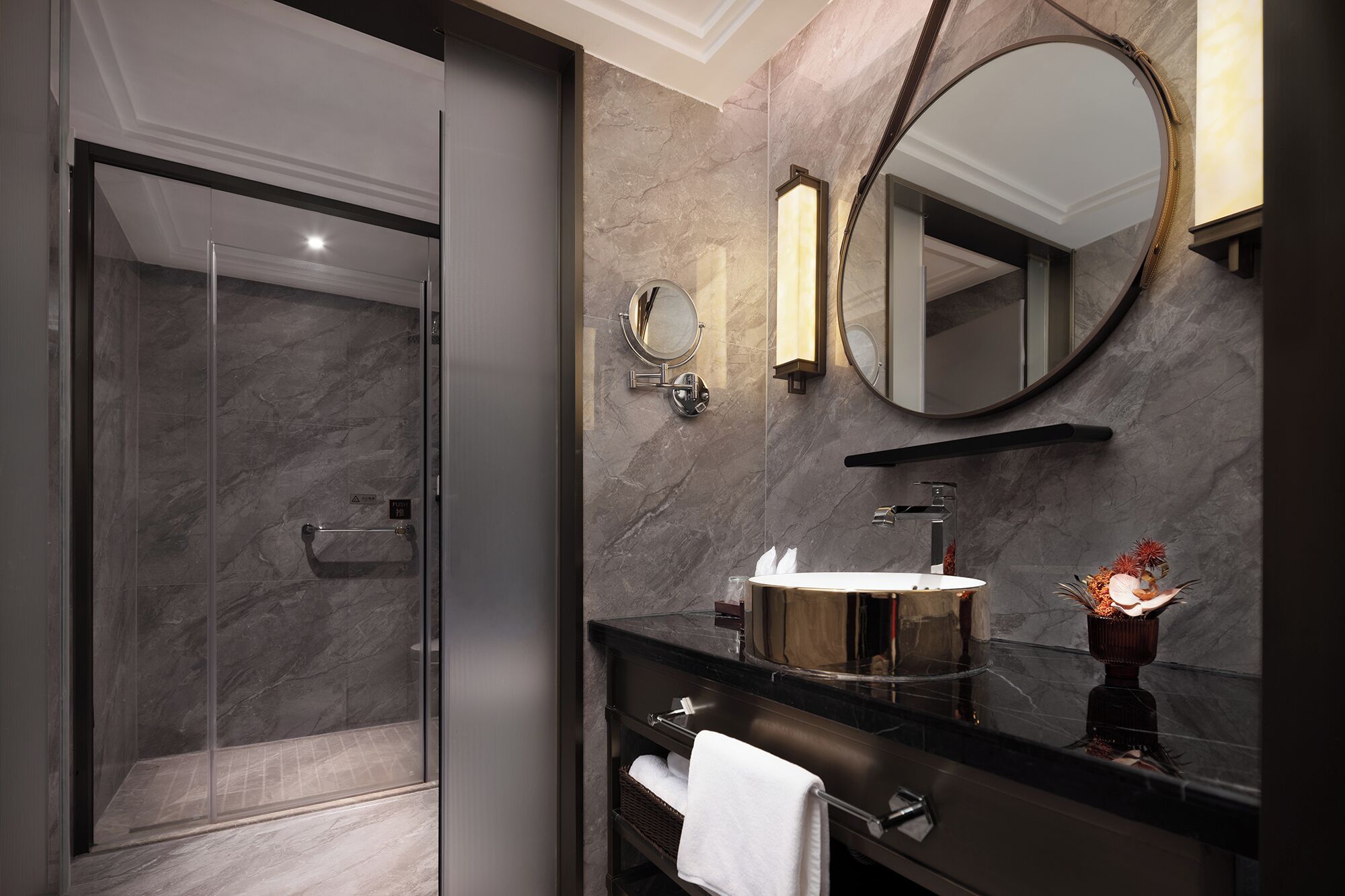 Un baño moderno con cabina de ducha de vidrio, pared con azulejos de mármol, espejo redondo y tocador con lavabo y perchero.