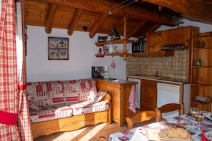 Une pièce chaleureuse avec un plafond en bois, un canapé agrémenté de coussins à motifs, un coin cuisine équipé d’un four à micro-ondes et d’un évier, ainsi qu’une table à manger avec une miche de pain et des verres à vin.