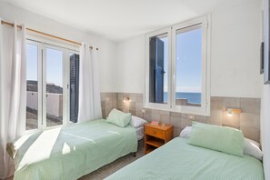 Una habitación con dos camas, una mesita de noche, una ventana con contraventanas y vistas al mar.