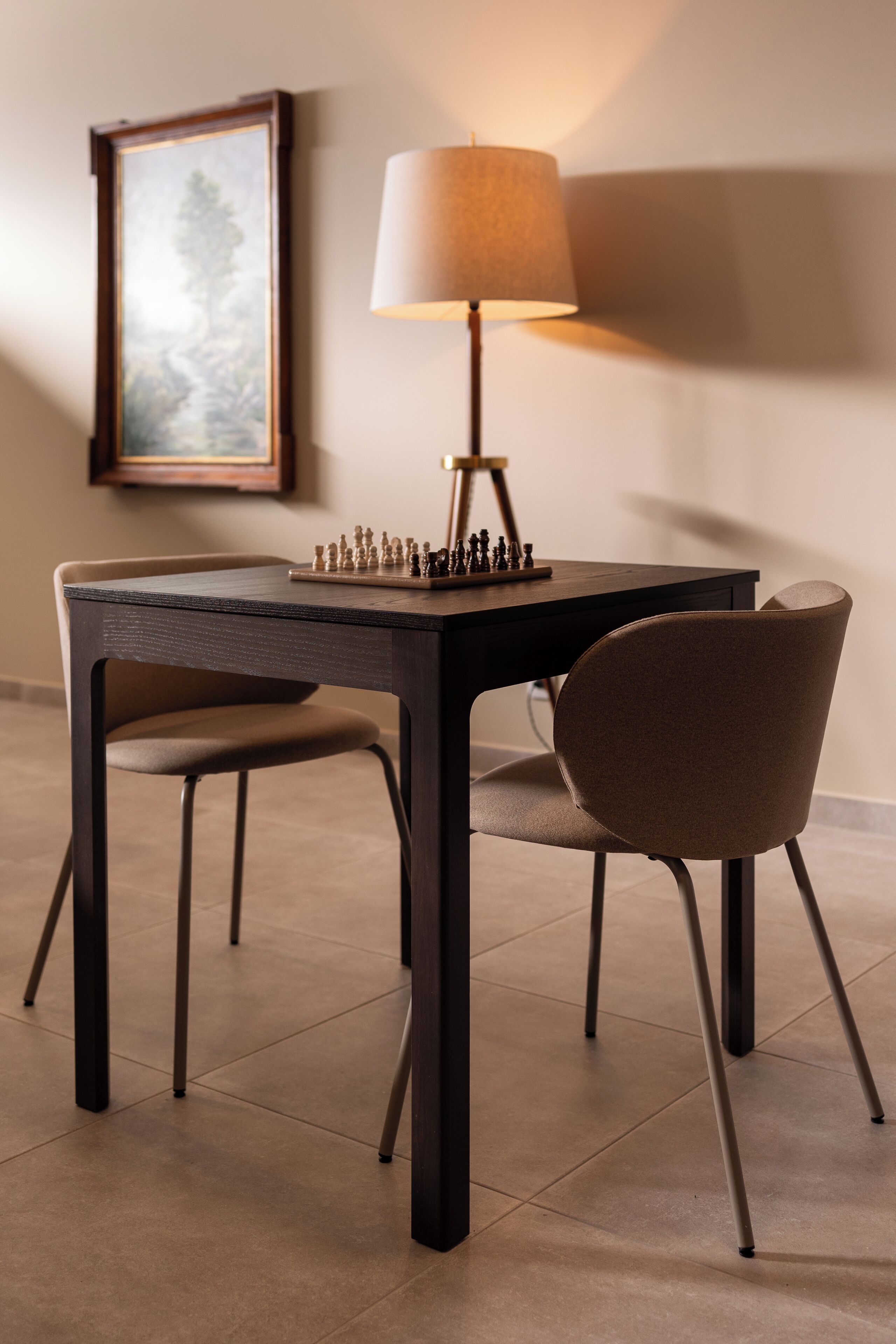 A dining table with a chess set, a lamp, and two chairs in a room with tiled flooring and a framed picture on the wall.