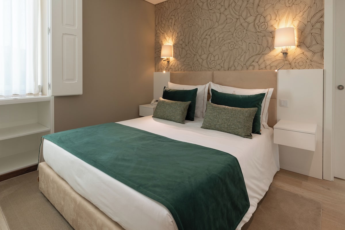 Una cama bien hecha con una colcha verde y almohadones, una mesita de noche con una lámpara, y una pared con estampado floral.