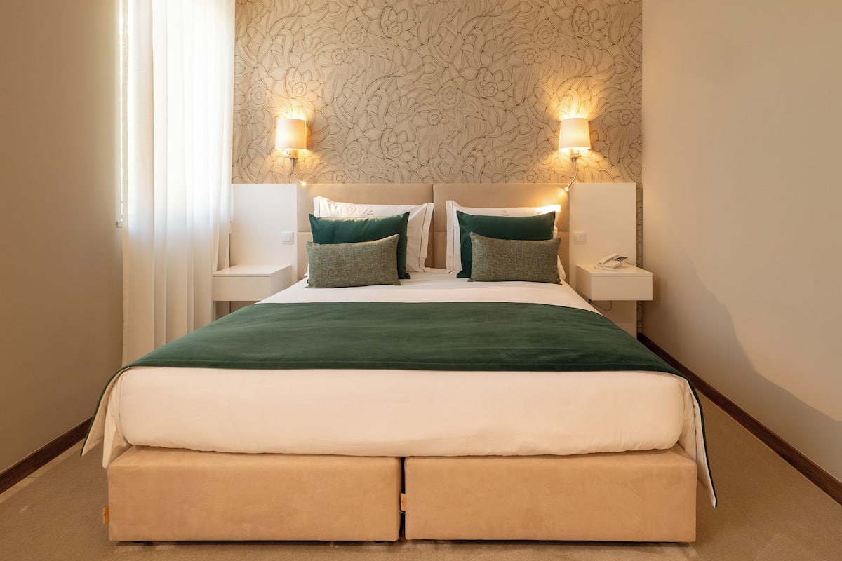 Una cama bien hecha con un edredón verde y dos almohadas, flanqueada por dos mesitas de noche con lámparas, en una habitación con papel tapiz estampado y piso de madera.