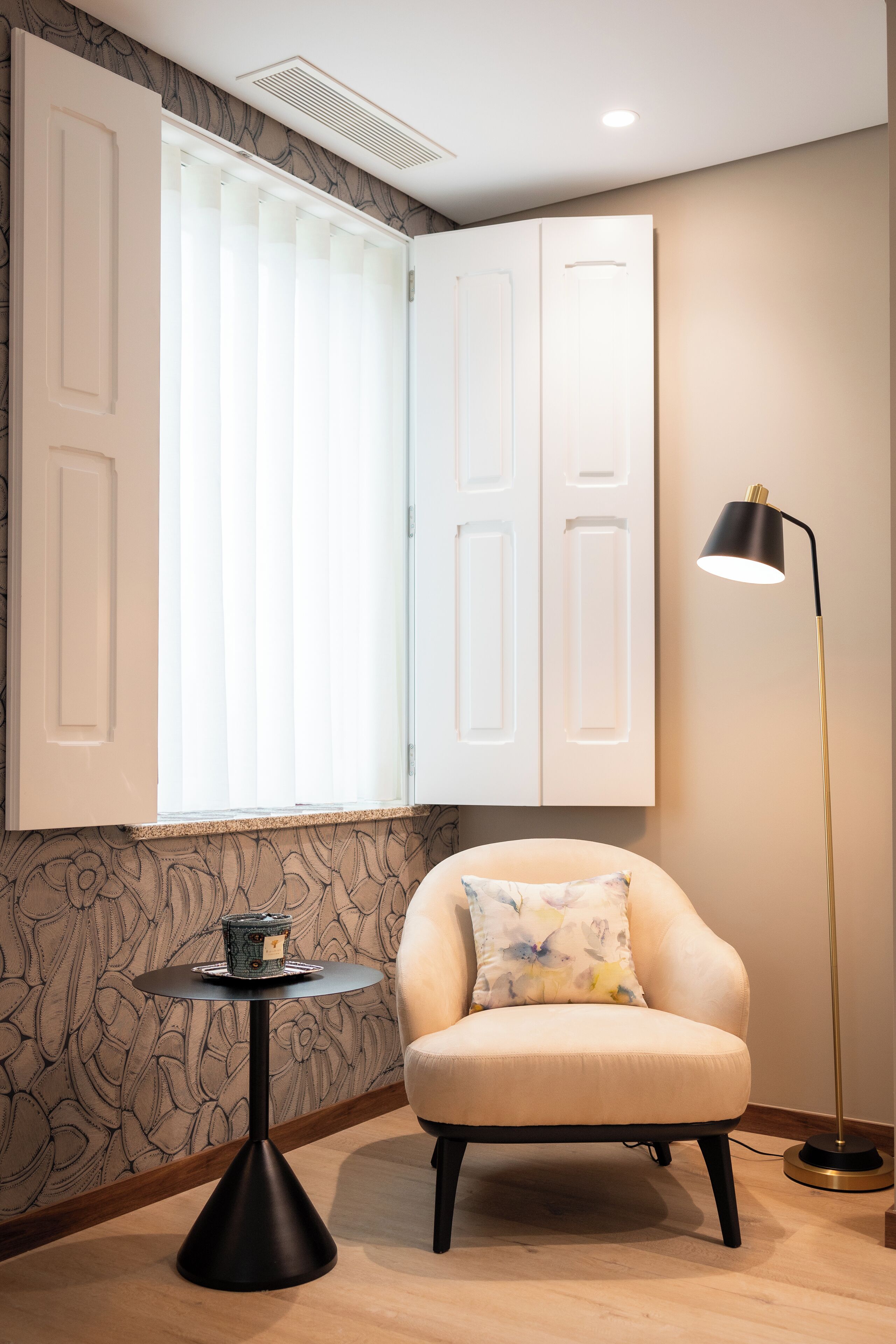 Une pièce avec un fauteuil blanc, une petite table ronde avec une bougie, un lampadaire et une fenêtre avec des stores.