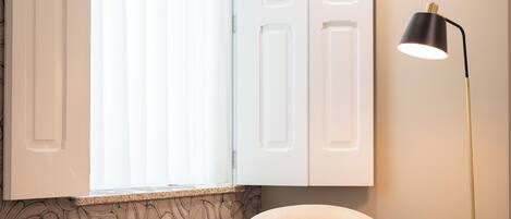 Una habitación con un sillón blanco, una mesita redonda con una vela, una lámpara de pie y una ventana con persianas.