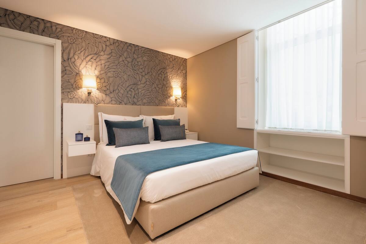 Un dormitorio con una cama, dos lámparas de noche, un ventanal con persianas y un papel tapiz estampado.