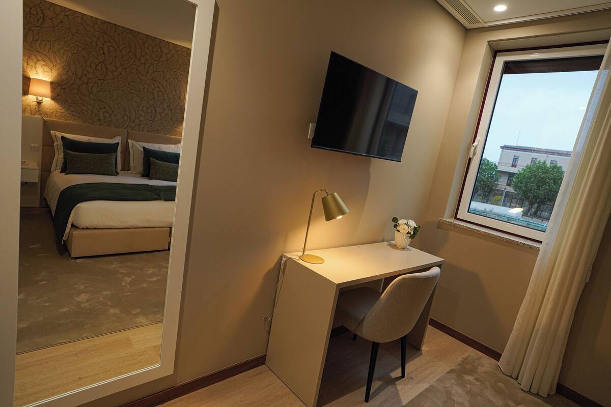 Habitación de hotel con cama, escritorio con silla, televisor montado en la pared y ventana con vista a edificios.
