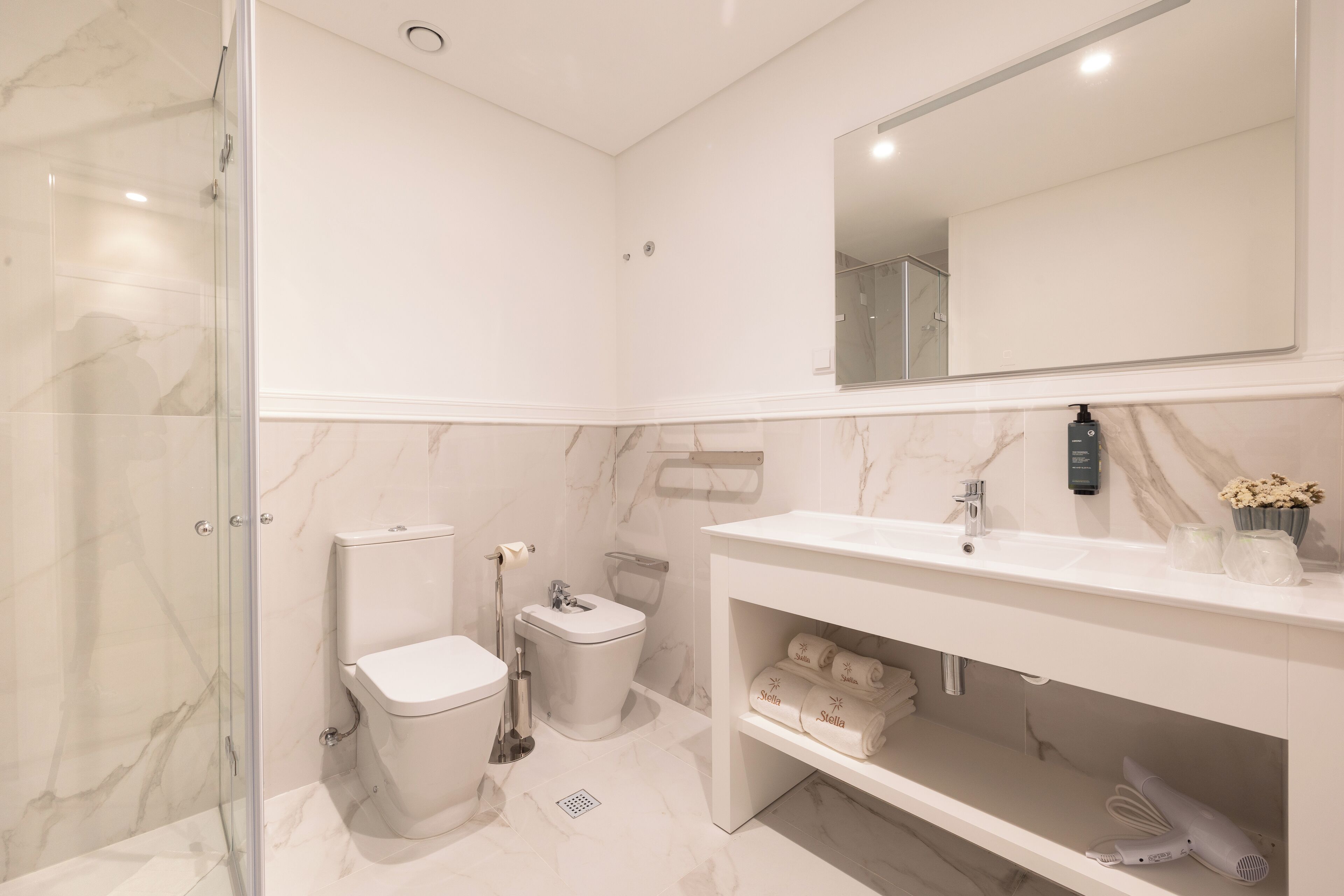Ein modernes Badezimmer mit Duschkabine aus Glas, einer weißen Toilette, einem Bidet, einem Waschtisch mit Spiegel und einem Regal mit Handtüchern und Fön.