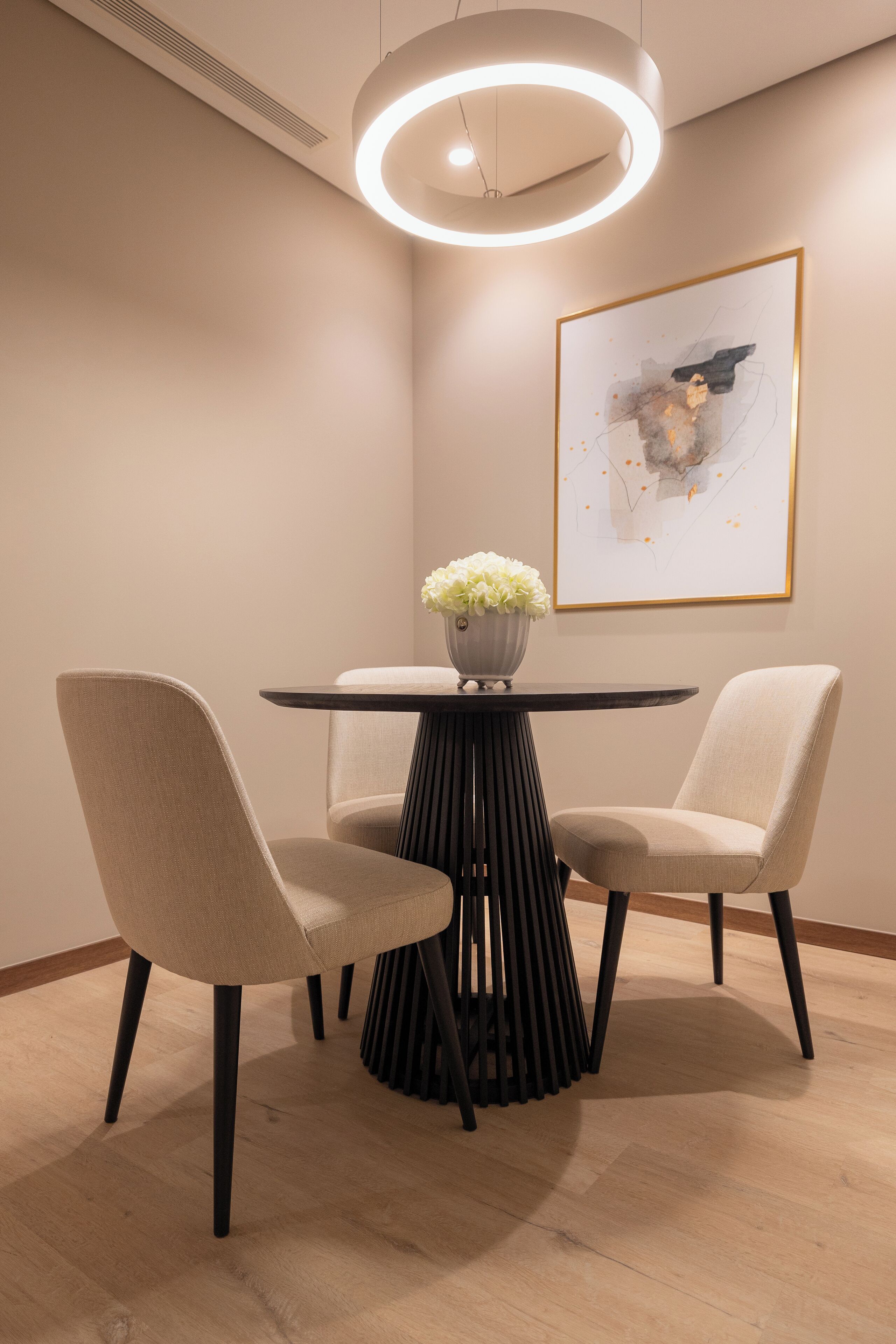 A modern dining area with a round table, two chairs, a vase with flowers, and a framed abstract painting on the wall.