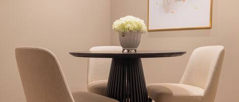A modern dining area with a round table, two chairs, a vase with flowers, and a framed abstract painting on the wall.
