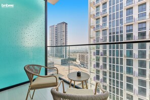 A balcony with wicker chairs and a small table, overlooking a cityscape with modern buildings. - bnbme - Binghatti Lavender - STUDIO (Dubai)
