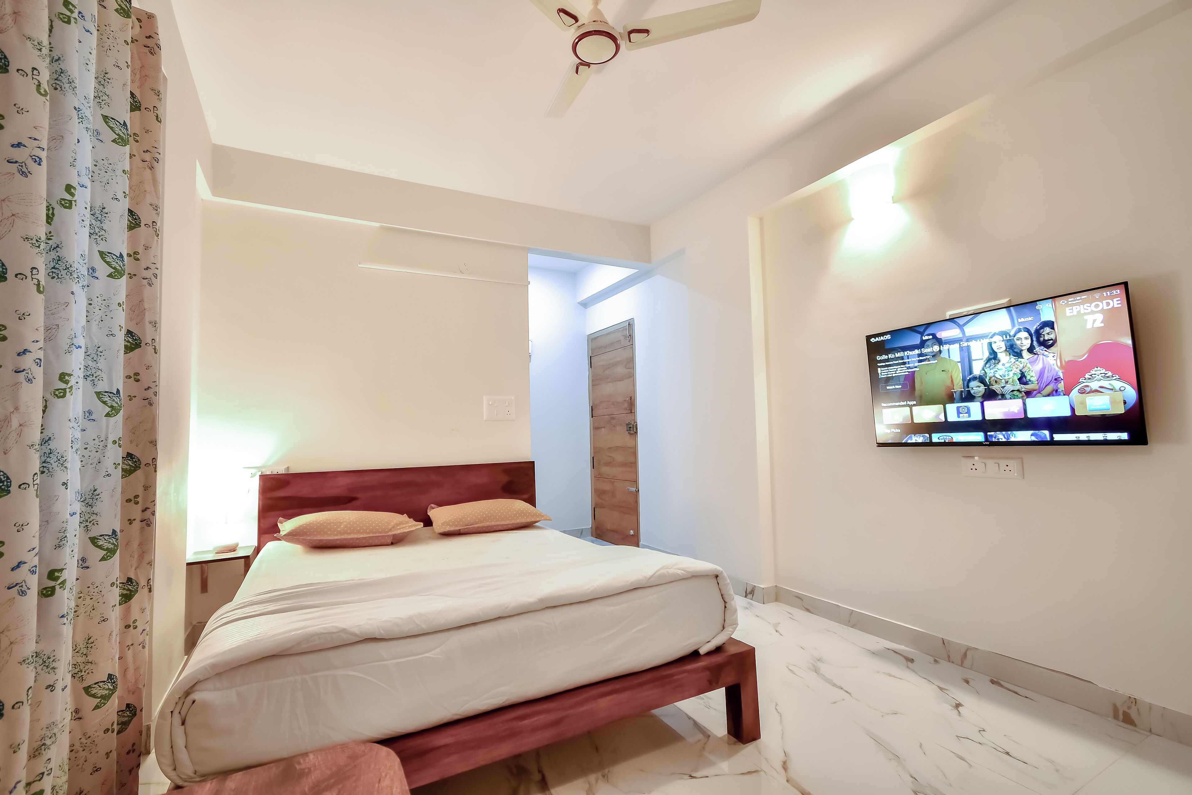A neatly made bed with a wooden headboard, a flat-screen TV mounted on the wall, and a ceiling fan.