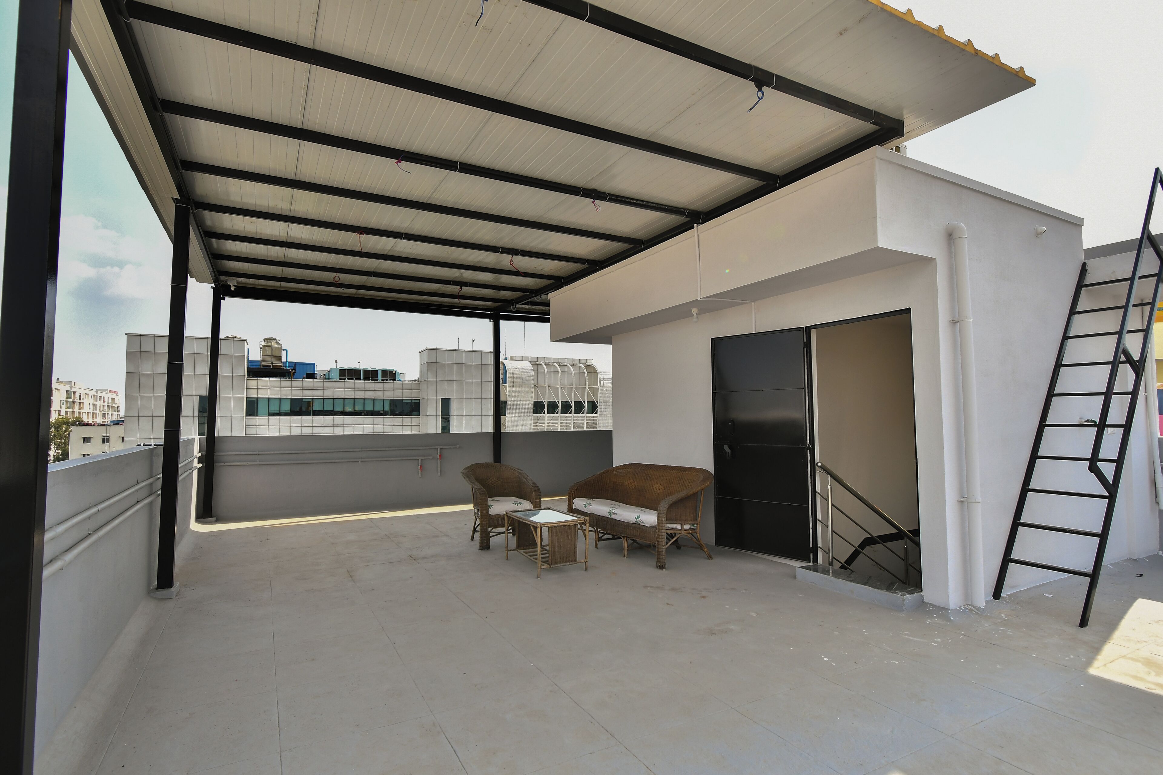 A covered outdoor area with two wicker chairs and a small table, a ladder, and a door leading to an upper floor.