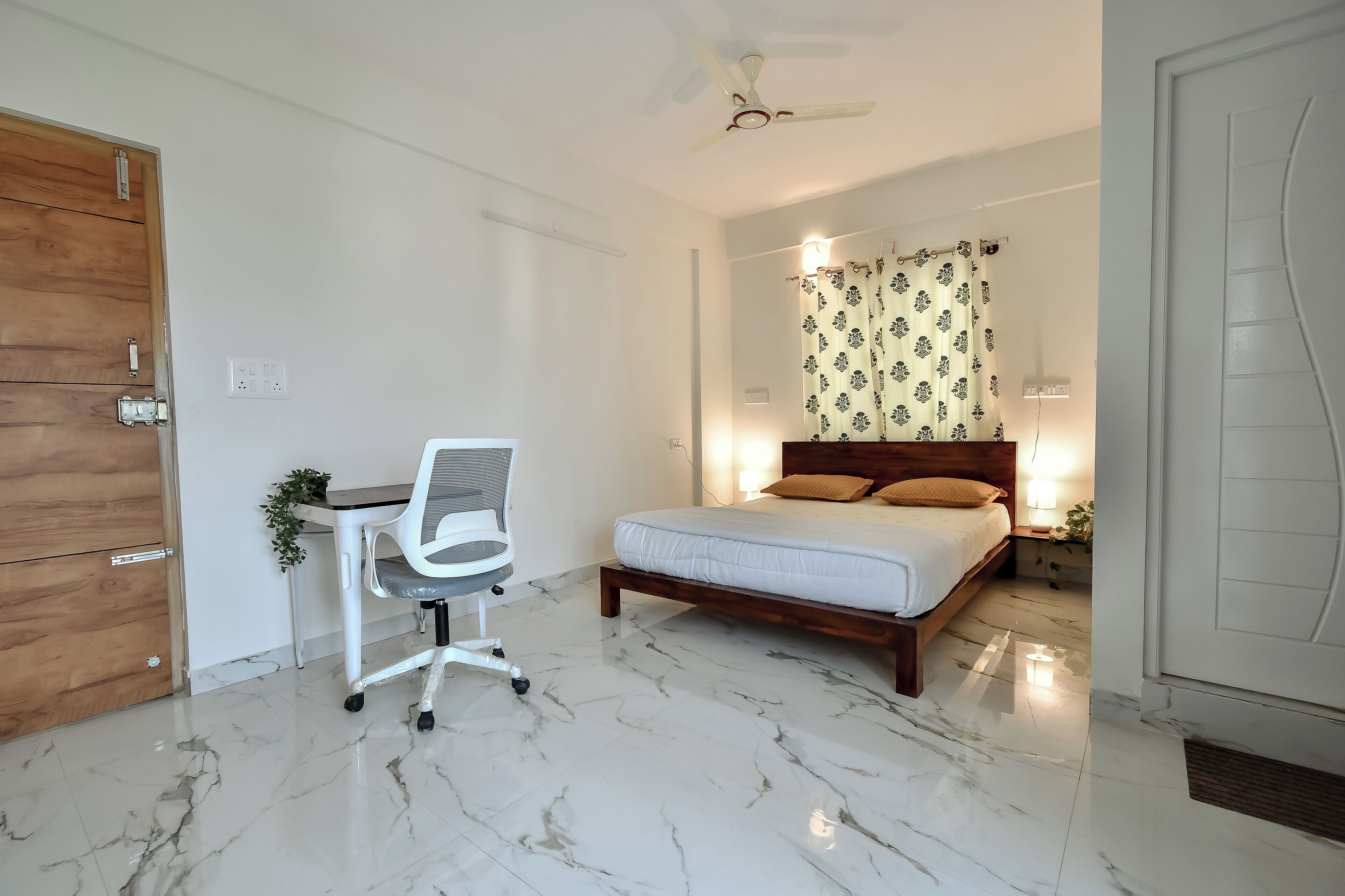 A bedroom with a bed, desk, chair, ceiling fan, and a nightstand with a lamp.