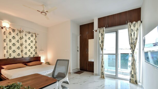 A modern bedroom with a bed, bedside table, chair, and a large window with curtains. - Namma HomeSlice (Bengaluru)