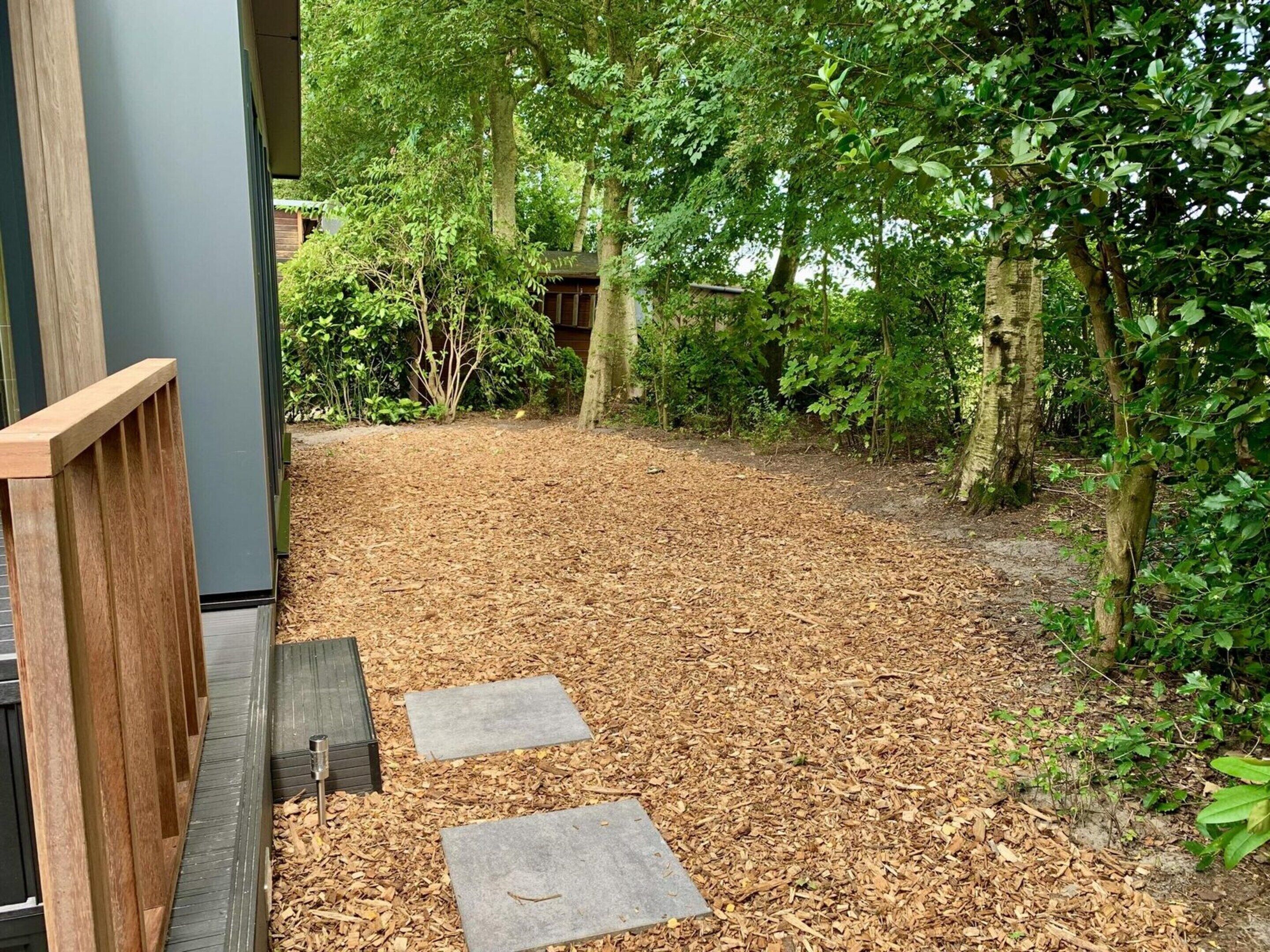 A pathway leading through a wooded area with a wooden fence on one side and a building on the other.