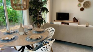 Un coin repas avec une table ronde dressée pour quatre personnes, entourée de chaises blanches ornées de motifs découpés. Une grande plante en pot se trouve à l’arrière-plan, et un téléviseur est fixé au mur au-dessus d’un meuble en bois.