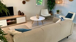 Un salon moderne avec un canapé beige, un fauteuil blanc, une petite table ronde et un téléviseur posé sur un meuble en bois.