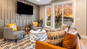 A living room with a flat-screen TV, patterned sofas, a coffee table, and large windows with a view of trees. - Monty by AvantStay | 4BDR 4BA | Balcony + Outdoor Seating | Minutes to Downtown (Nashville)