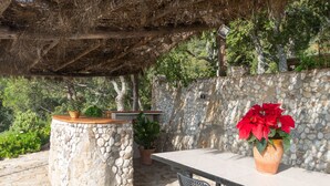 Eine Terrasse im Freien mit einem Steintisch und -stühlen, umgeben von einer Steinmauer und einem Reetdach.