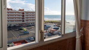 Blick aus einem Hotelzimmerfenster auf einen Parkplatz, einen Strand und ein Gebäude mit Balkonen.