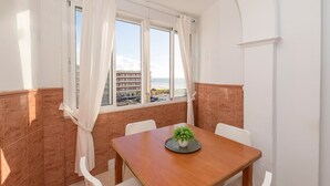 Ein Essbereich mit einem Holztisch und weißen Stühlen, einer Topfpflanze auf dem Tisch und Blick auf den Strand und Gebäude durch große Fenster.