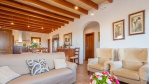 A spacious living room with a beige sofa, two armchairs, and a coffee table with a vase of flowers. The room features wooden beams on the ceiling and a kitchen area in the background. - Son Morro Ca Na Poveta (Santa Margalida)