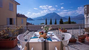 A balcony with a table set for two, overlooking a lake and mountains. - Fontana Del Lago Apt 7 (Varenna)