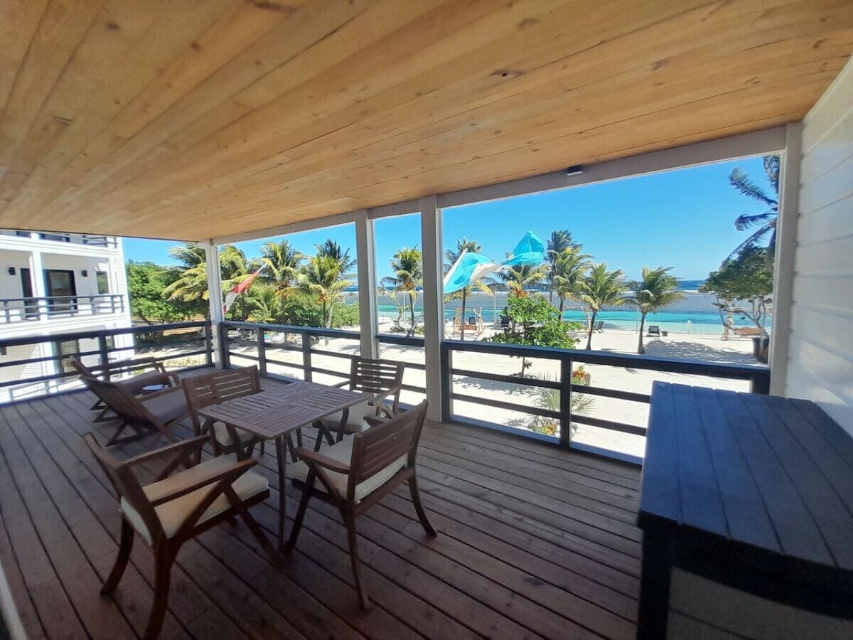 A wooden deck with a table and chairs, overlooking a beach with palm trees and umbrellas.