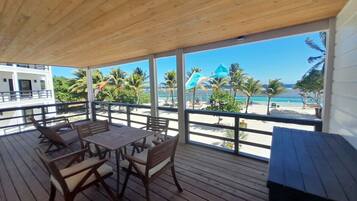 A wooden deck with a table and chairs, overlooking a beach with palm trees and umbrellas.