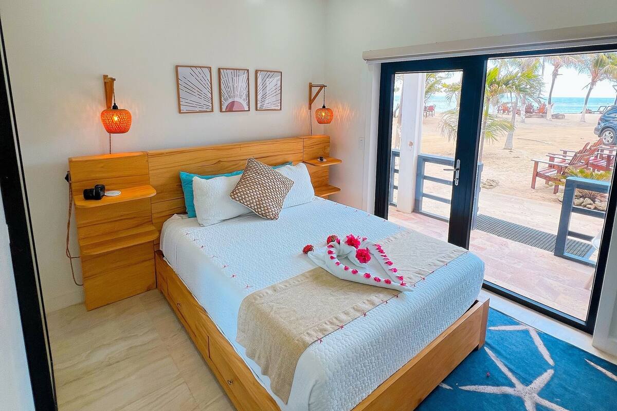 A bedroom with a bed, wooden headboard, bedside tables, a ceiling fan, and a view of the beach through an open door.