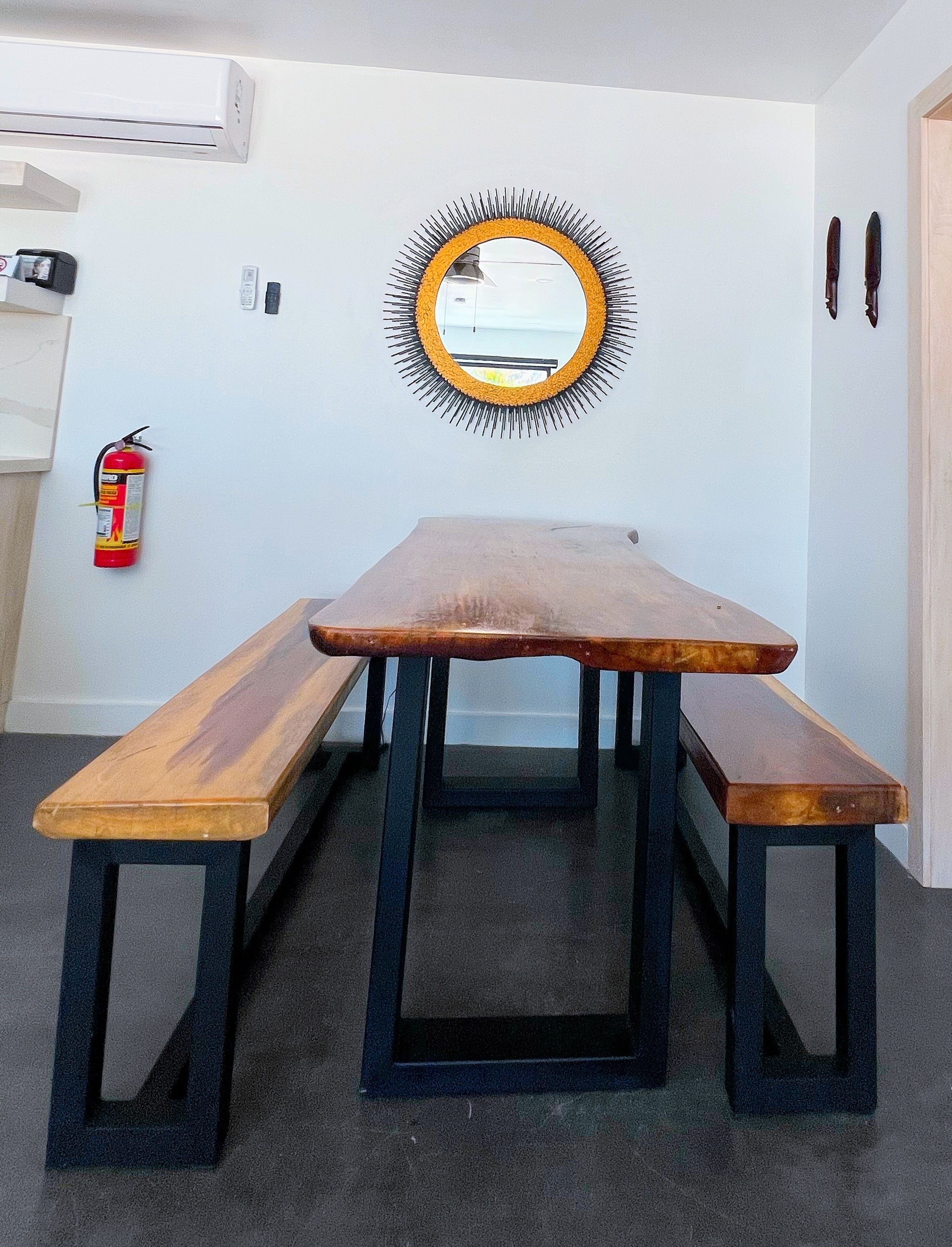 Una mesa de madera con bancos, un espejo con marco de sol, un extintor y un equipo de aire acondicionado instalado en la pared.