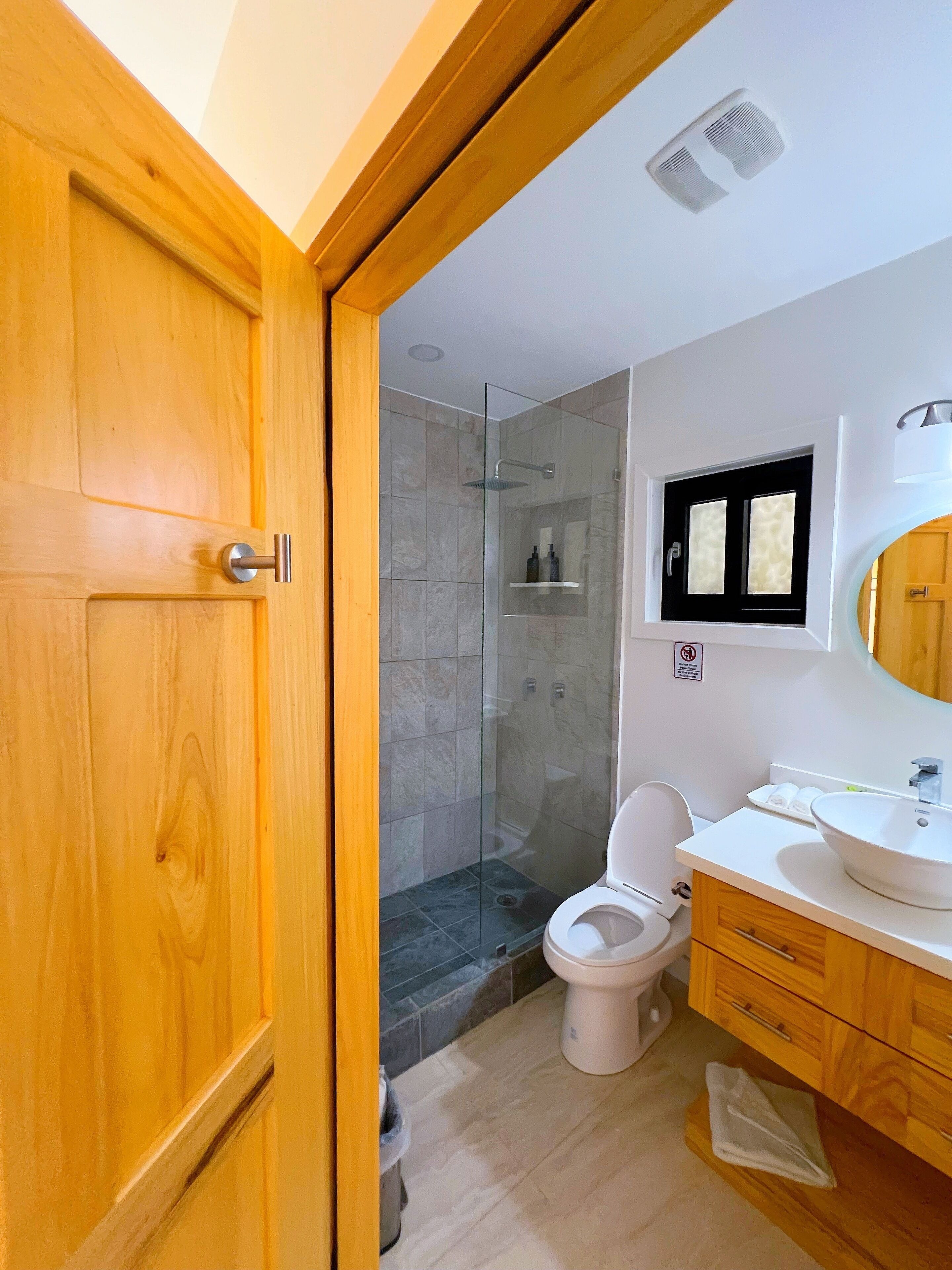 Ein modernes Badezimmer mit Holztür, Duschkabine aus Glas, weißer Toilette und Waschtisch mit Spiegel.